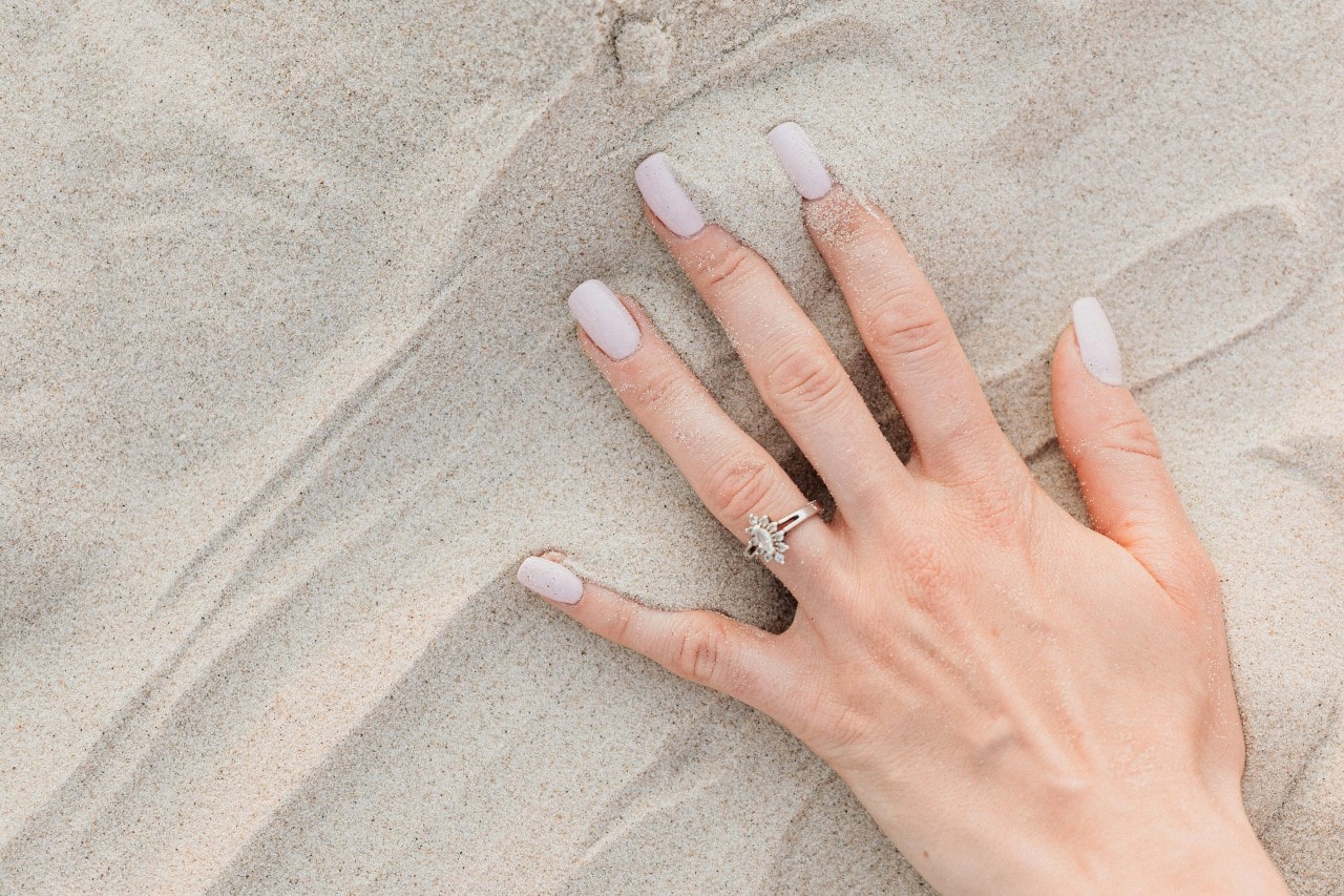 A woman's hand in the sand, wearing a white gold engagement ring A woman's hand in the sand, wearing a white gold engagement ring