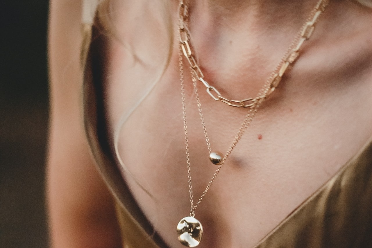 Close-up of a woman wearing layered gold necklaces; one with a chain-link design, the other with small gold pendants, against a satin garment. Close-up of a woman wearing layered gold necklaces; one with a chain-link design, the other with small gold pendants, against a satin garment.