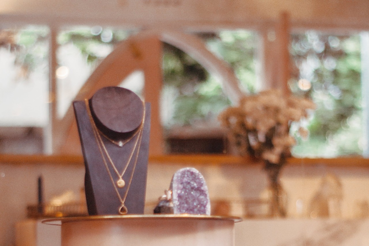 A display of layered gold necklaces on a black bust in a softly lit jewelry store, with a bouquet of flowers and a purple geode in the background. A display of layered gold necklaces on a black bust in a softly lit jewelry store, with a bouquet of flowers and a purple geode in the background.