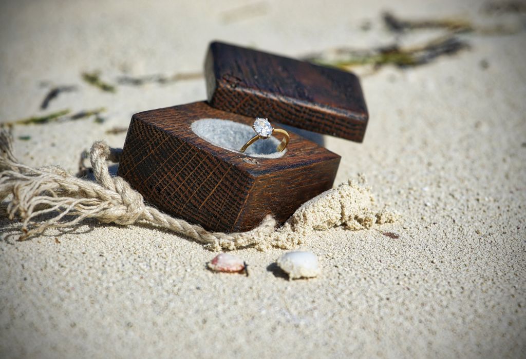 A gold oval-cut engagement ring sits in a wooden box in the sand A gold oval-cut engagement ring sits in a wooden box in the sand