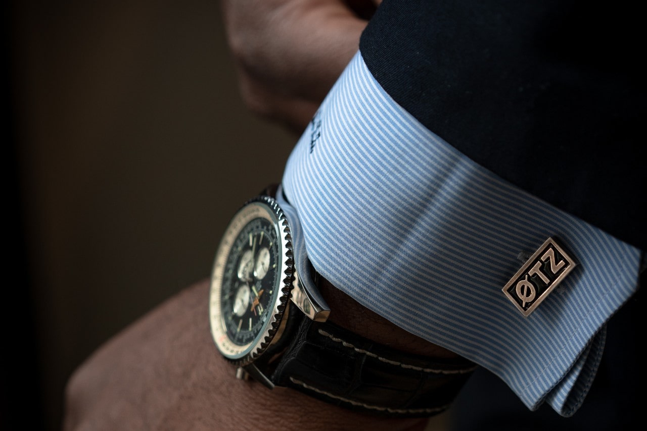 A close-up of a man's wrist wearing a luxury chronograph watch with a black leather strap and a blue shirt with custom cufflinks. A close-up of a man's wrist wearing a luxury chronograph watch with a black leather strap and a blue shirt with custom cufflinks.
