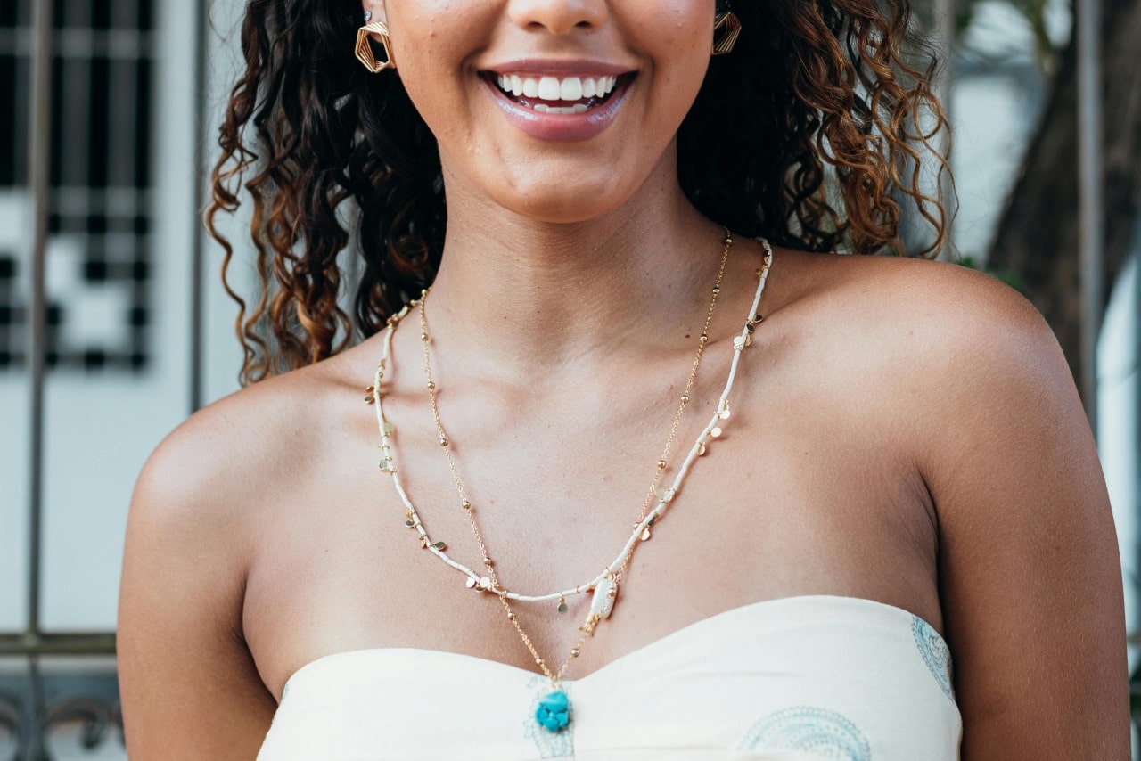A close-up of a woman smiling in a white strapless top, showcasing a layered yellow gold necklace with a blue stone in the center. A close-up of a woman smiling in a white strapless top, showcasing a layered yellow gold necklace with a blue stone in the center.