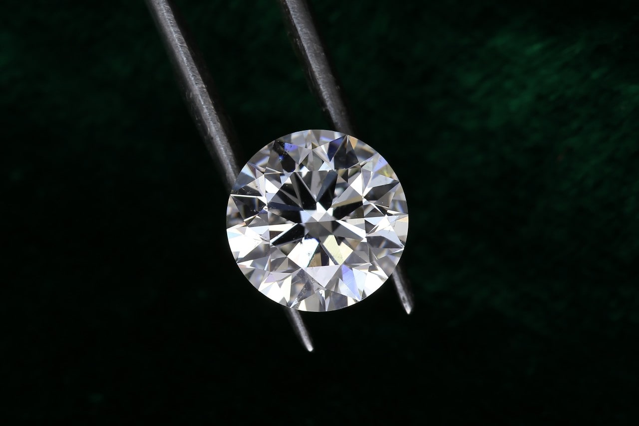A professional jeweller holds a round-cut lab-created diamond with a pair of jewellery tweezers against a dark green background A professional jeweller holds a round-cut lab-created diamond with a pair of jewellery tweezers against a dark green background