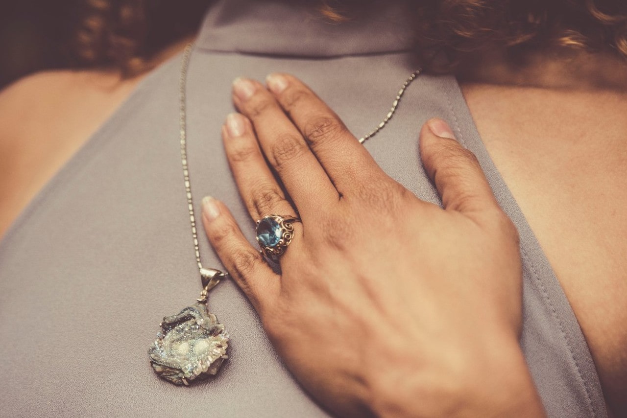 A close-up of a woman with her hand on her heart, wearing an elegant necklace and bold ring.