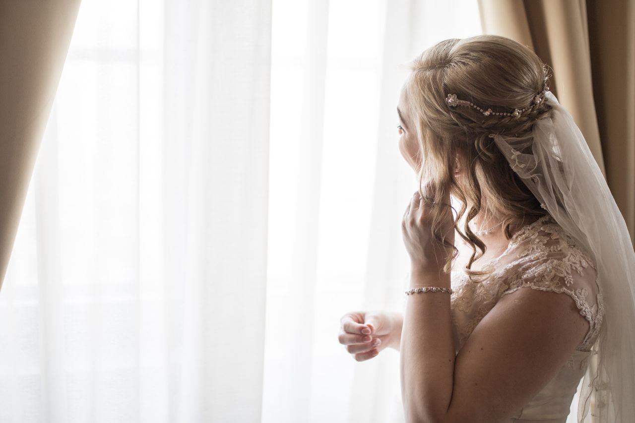 A close-up of a bride-to-be putting jewellery on as she gazes out a brightly lit window.