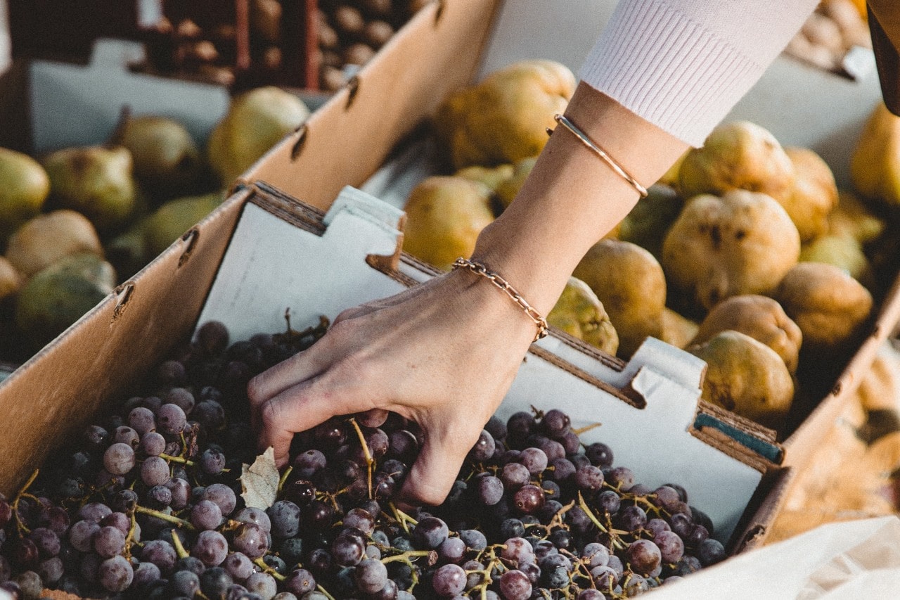 A close-up of a woman’s hand adorned with bracelets, selecting fresh grapes at a farmer’s market.