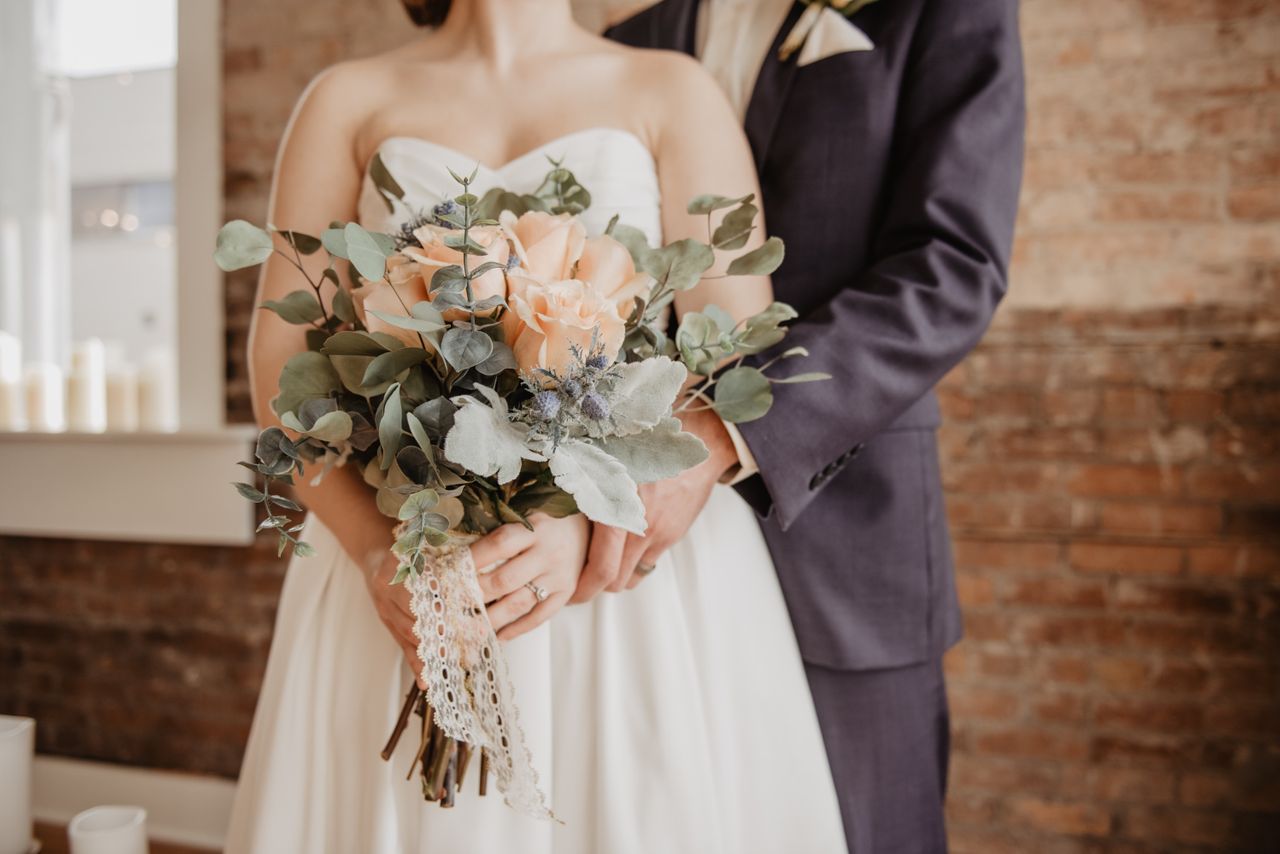 A close-up of a groom embracing his bride from behind as she holds an elegant bouquet.