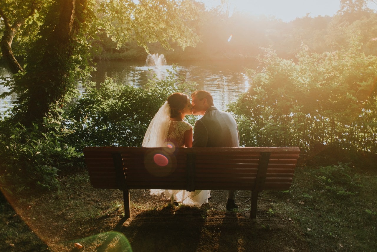 A newlywed couple sharing a kiss on a bench overlooking an enchanting lake with a fountain.