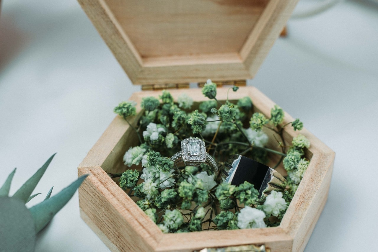 A stunning diamond engagement ring inside a wooden box, surrounded by small flower buds.