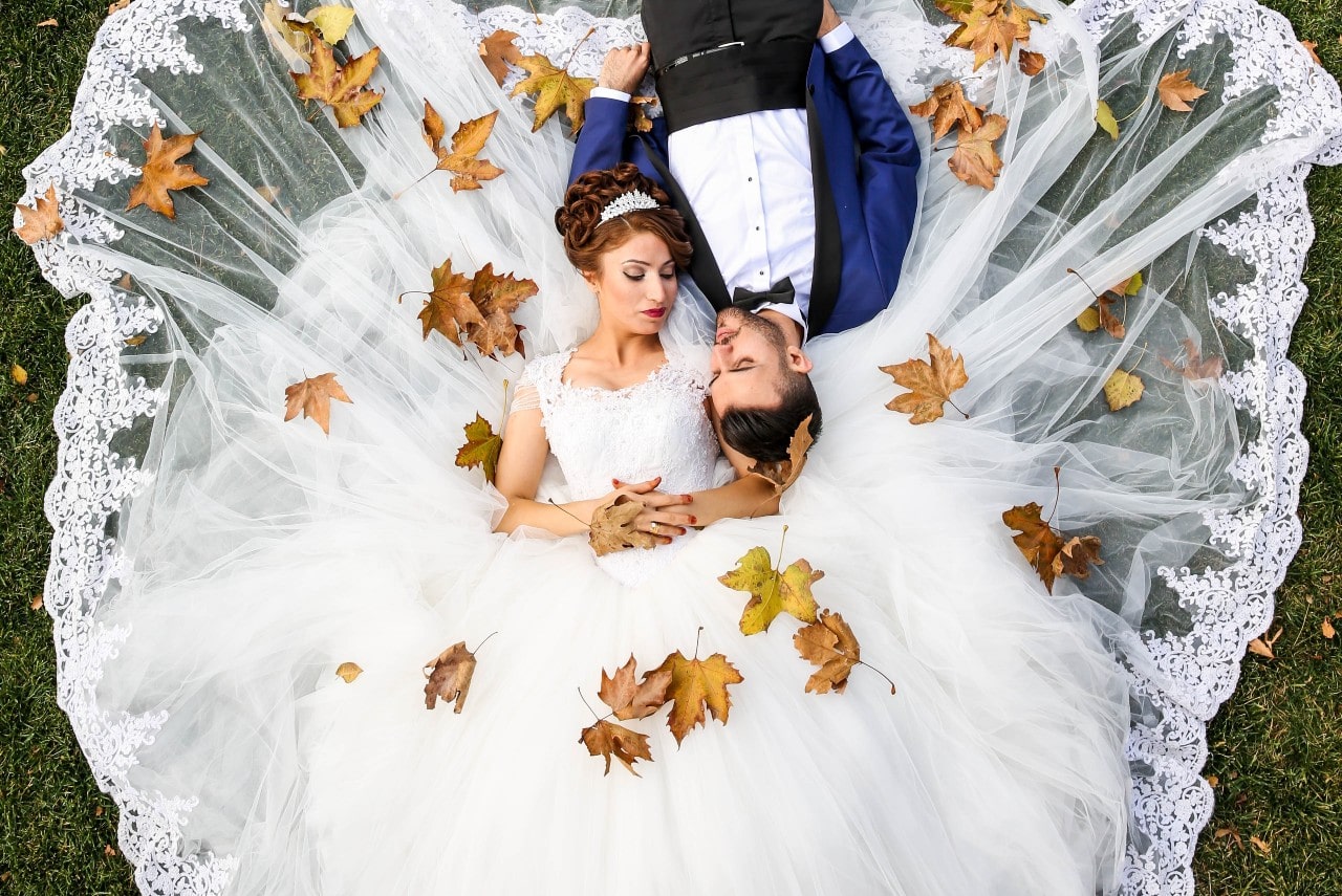 An overhead photo of a bride and groom laying in the grass, among autumn leaves.