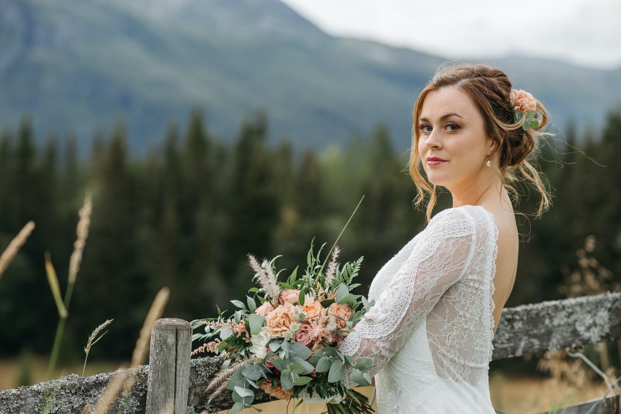 A bride standing before a rustic wooden fence in the mountains, holding an elegant bouquet.