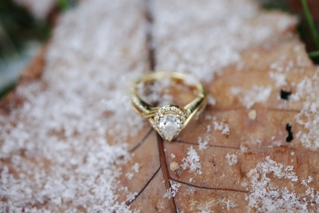 A close-up of an elegant halo engagement ring with a pear center stone, displayed on a snowy winter leaf.